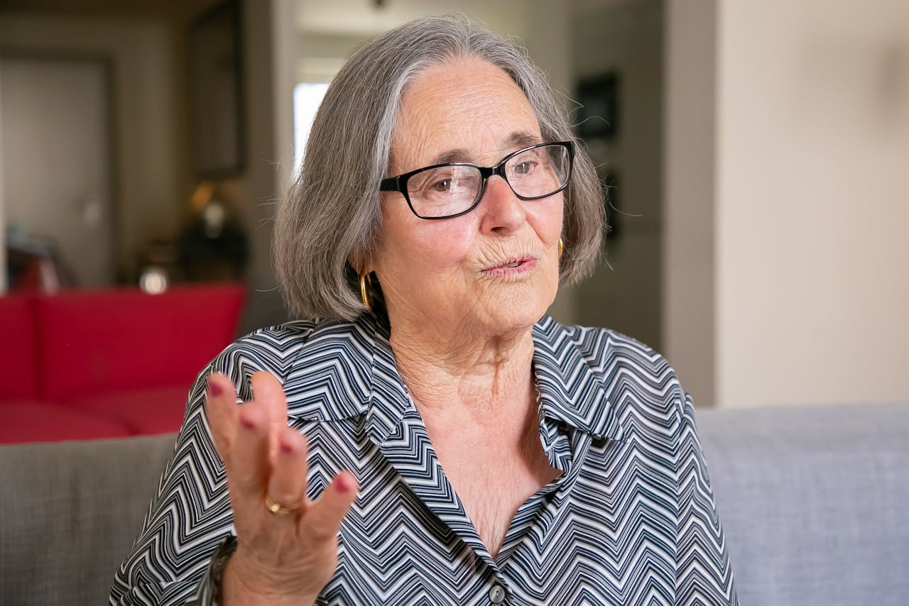 A senior caucasian woman with glasses speaking animatedly indoors.